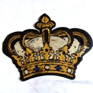 May include: A crown-shaped patch featuring gold, silver, and black sequins. The design includes a central fleur-de-lis element, a band decorated with small blue and white circular accents, and a solid black border outlining the entire shape.