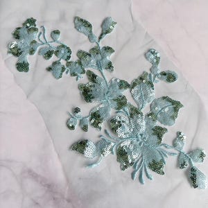 May include: A light blue floral applique patch featuring intricate embroidery and shimmering sequins on a sheer mesh fabric base. The design consists of delicate stems, leaves, and flower blossoms in a soft blue hue with metallic green accents.