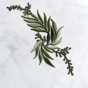 Puede incluir: Un parche bordado con un diseño botánico de hojas verde salvia y delicadas ramitas con pequeños brotes. El bordado utiliza varios tonos de hilo verde para crear profundidad y textura en el follaje, dispuesto en un patrón curvo y fluido.