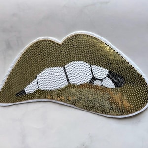 Könnte beinhalten: Ein goldfarbener Pailletten-Lippen-Aufnäher mit weißen und schwarzen Details. Die Lippen sind weiß umrandet und haben ein weißes Perlendesign für die Zähne. Der Aufnäher ist ein dekorativer Artikel, wahrscheinlich für Kleidung oder Accessoires.