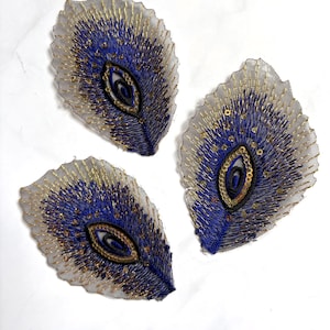 Può includere: Tre toppe ricamate a forma di piuma di pavone con fili blu e oro su una base bianca trasparente. Ogni toppa presenta un motivo centrale a occhio accentuato da paillettes dorate e sottili cuciture che si irradiano a forma di foglia.