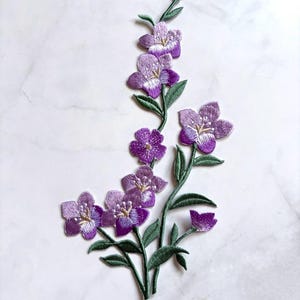 May include: An embroidered patch featuring a vertical arrangement of purple flowers with white centers and green stems and leaves. The design includes multiple blossoms in varying shades of violet, with detailed stitching throughout the petals and foliage.