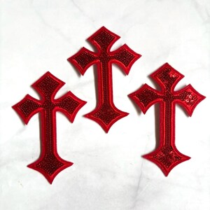 May include: Three identical cross-shaped patches in a vibrant red color. Each patch features a central cross design filled with small, reflective red sequins and is outlined with a smooth, solid red fabric border, creating a textured appearance.