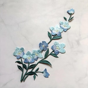 May include: Embroidered floral applique featuring light blue flowers with darker blue and purple accents. The flowers are attached to green stems with green leaves. The applique is on a white marble background.
