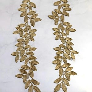 May include: Two gold-colored leaf branch appliques. Each applique features a detailed design of leaves and stems, with a metallic sheen. The appliques are arranged on a white marble surface, creating a contrast between the gold and white.