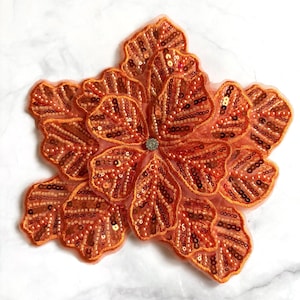 Könnte beinhalten: Eine dekorative orangefarbene Blumenapplikation mit geschichteten Blütenblättern, die mit passendem Garn bestickt und mit schimmernden Pailletten sowie kleinen Perlen verziert sind. Ein einzelner klarer Strassstein sitzt in der Mitte.