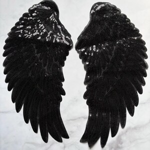 Puede incluir: Dos parches de alas de ángel negros con lentejuelas. Cada ala está detallada con filas de lentejuelas negras y una dispersión de lentejuelas plateadas. Las alas se muestran sobre una superficie de mármol blanco.