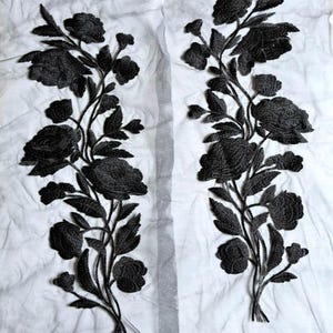 Puede incluir: Dos aplicaciones de bordado floral negro dispuestas verticalmente con rosas, capullos y hojas detalladas. El intrincado trabajo de hilo negro está cosido sobre una base de tela de malla blanca, fina y transparente, creando un diseño de alto contraste.