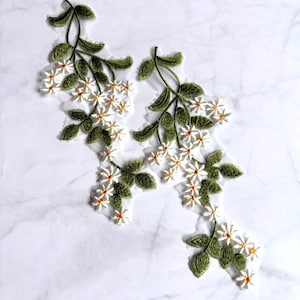 Puede incluir: Dos apliques bordados florales con flores blancas similares a las margaritas, con centros amarillos y hojas verdes. El diseño botánico está cosido sobre una base de malla transparente, formando una delicada disposición de enredadera.