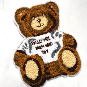 May include: A brown teddy bear patch with a white t-shirt that has the text "DOLLS NOT MAKE ME TOY". The bear has a light brown belly and paws, and a dark brown outline. The patch is made of a textured material.