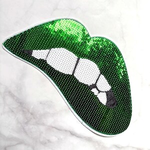 May include: A green sequin lips patch with white teeth and black detailing. The patch has a white border and is shaped like a pair of lips. The sequins give the patch a shiny, textured look.