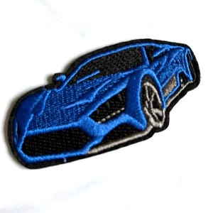 May include: An embroidered patch featuring a stylized blue sports car with black detailing and silver-colored wheels. The design is outlined in black thread, showcasing the sleek, aerodynamic silhouette of the vehicle from a three-quarter angle.