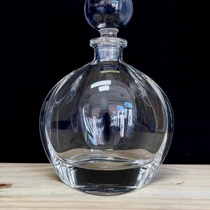 May include: A clear glass decanter with a round body and a spherical stopper. The decanter has a label that reads "CRYSTALITE BOHEMIA". The glass reflects the surrounding environment. The decanter sits on a wooden surface against a black background.