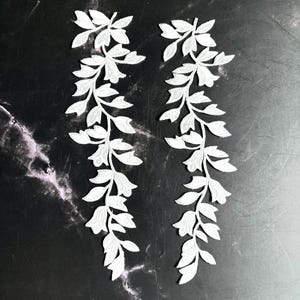 Könnte beinhalten: Zwei identische weiße Stickapplikationen mit einem detaillierten botanischen Rankenmuster aus mehreren Blättern und kleinen Blütenknospen. Die Stickerei ist dicht und strukturiert und zeigt eine symmetrische Anordnung von Blattwerk.