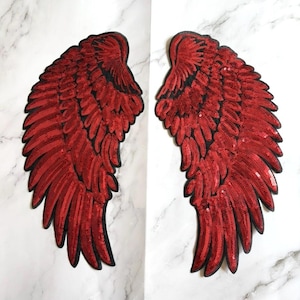 May include: Pair of red sequined angel wings with black trim. The wings are symmetrical, with each wing featuring a detailed feather design. The sequins give the wings a shimmering effect, perfect for adding a touch of glamour to any project.