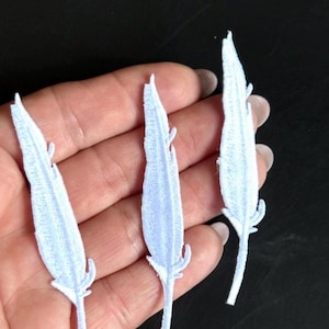 Peut inclure: Trois écussons brodés en forme de plume blanche avec des coutures détaillées sur les bords. Chaque écusson présente une pointe effilée et une tige fine à la base, mettant en valeur une surface texturée créée par le travail dense du fil.