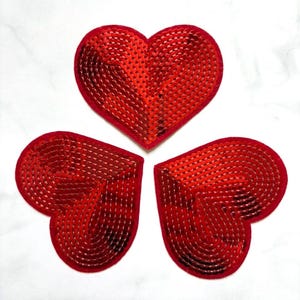 Può includere: Tre toppe rosse a forma di cuore con superficie di paillettes. I cuori sono disposti a trifoglio su sfondo bianco. Le toppe hanno un bordo in feltro.