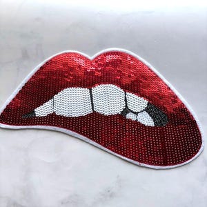Könnte beinhalten: Ein roter Lippen-Aufnäher mit Pailletten, weißen Zähnen und einem schwarzen Akzent. Der Aufnäher ist weiß umrandet und dient wahrscheinlich dekorativen Zwecken, wie z. B. zum Verschönern von Kleidung oder Accessoires. Die Pailletten verleihen dem Aufnäher ein glänzendes, strukturiertes Aussehen.