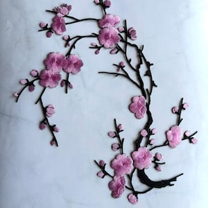 May include: Embroidered floral applique featuring a branch with pink cherry blossoms and buds. The blossoms have a detailed, textured appearance, and the branches are black. Ideal for fabric embellishment or craft projects.