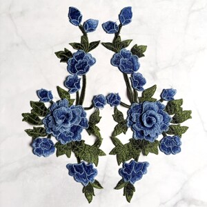May include: Two symmetrical floral embroidery patches featuring detailed blue roses and buds with olive green leaves. The intricate stitching creates a textured appearance for these decorative appliqués, which are designed to be placed together.