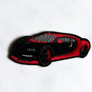 May include: An embroidered patch featuring a sleek sports car in black and red thread. The design includes silver-toned stitching for the windshield and windows, with a black border outlining the vehicle's aerodynamic shape and low-profile wheels.