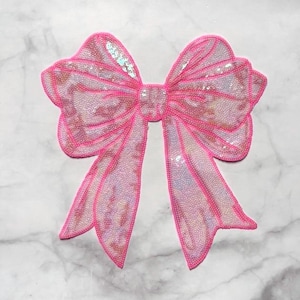 Op de afbeelding: Een grote, roze paillettenstrik met een felroze omtrek. De strik heeft een klassieke vorm met twee lussen en twee linten. De pailletten vangen het licht op en creëren een glinsterend effect. De strik is waarschijnlijk bedoeld om te knutselen of te verfraaien.