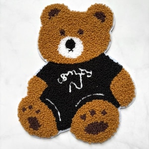 Puede incluir: Una alfombra con forma de oso de peluche marrón con una camiseta negra con texto blanco en cursiva. El oso tiene una cara blanca, patas marrones y un aspecto texturizado y esponjoso. La alfombra es probablemente hecha a mano.