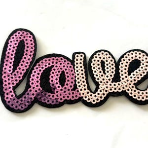 May include: A decorative patch featuring the word Love in cursive script. The letters are covered in sequins, with the L and o in a vibrant pink gradient and the v and e in a soft pale pink. The entire word is outlined on a black felt backing.