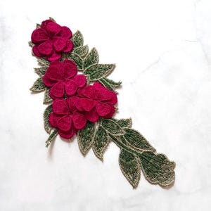May include: A floral applique featuring three magenta flowers and green leaves with gold-colored accents. The embroidered design is set against a white background, showcasing the intricate details of the fabric.