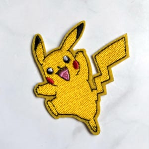 Puede incluir: Un parche bordado que muestra un personaje amarillo con orejas largas de puntas negras, mejillas circulares rojas y una boca abierta que muestra una lengua rosa. El personaje tiene una cola dentada en forma de rayo y una pose dinámica.