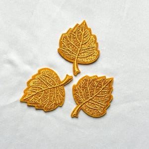 May include: Three embroidered patches shaped like leaves in a vibrant golden yellow colour. Each patch features detailed stitching that mimics the veins of a leaf, with a distinct stem extending from the base of each individual leaf shape.