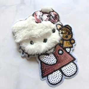 Puede incluir: Un parche decorativo con la cara de un gato de peluche blanco con un lazo de lentejuelas rosa. El gato lleva un vestido de lentejuelas rojo y sostiene un pequeño oso de peluche marrón. El parche está delineado en negro.