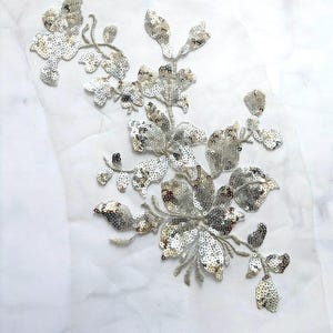 Könnte beinhalten: Blumenapplikation mit silbernen Pailletten. Das Design zeigt Blumen und Blätter, die alle mit schimmernden Pailletten bedeckt sind. Die Applikation ist auf einem transparenten, weißen Stoffrücken befestigt, was einen zarten und eleganten Look erzeugt. Ideal zum Verzieren von Kleidungsstücken oder Accessoires.