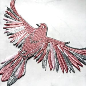 May include: Embroidered bird patch in flight, with pink, silver, and white thread detailing. The bird's wings are outstretched, displaying the intricate stitching and colour gradients. Suitable for clothing or accessories, this patch is a decorative item.