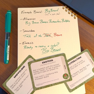 May include: A notebook with green ink notes, including "Big Brown!" and examples of literary devices. A teal pen and three wordplay cards are on a wooden surface. The notebook has a spiral binding.
