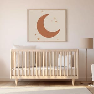 Peut inclure: Une affiche encadrée représentant une lune en croissant et des étoiles terracotta sur fond crème. L'œuvre est au-dessus d'un lit bébé en bois clair avec des oreillers blancs. Un ours en peluche est sur une petite chaise.