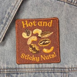 Puede incluir: Un chaleco de mezclilla azul claro con un parche cuadrado bordado en el pecho. El parche es marrón con texto amarillo que dice Hot and Sticky Nuts! y representa varias nueces, incluyendo anacardos, nueces y semillas de girasol en el centro.