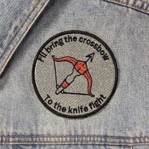 Op de afbeelding: Een lichtblauw spijkergilet met een ronde geborduurde patch op de borst. De patch heeft een grijze achtergrond, een rode kruisboogillustratie en de zwarte tekst "I'll bring the crossbow To the knife fight."