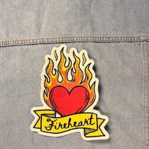 May include: A light blue denim jacket featuring a large embroidered patch on the back. The patch depicts a red heart surrounded by yellow and orange flames, positioned above a yellow banner with the black cursive text Fireheart written on it.