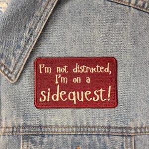 Könnte beinhalten: Eine hellblaue Jeansweste mit Kragen, Metallknöpfen und einem rechteckigen gestickten Aufnäher. Der Aufnäher ist dunkelrot mit weißem Text, der lautet: I'm not distracted, I'm on a sidequest! Die Weste hat ein Etikett mit der Aufschrift L.A. BLUES.