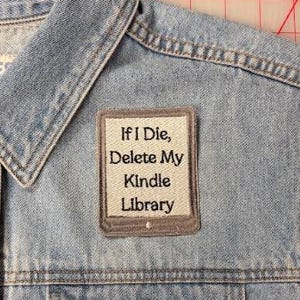 Op de afbeelding: Een lichtblauw spijkergilet met een rechthoekige geborduurde patch op de borst. De patch heeft een lichtgrijze rand en een crèmekleurig midden met de zwarte geborduurde tekst: If I Die, Delete My Kindle Library. Het gilet heeft metalen knopen.