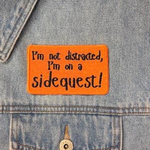 May include: A rectangular orange embroidered patch with black text that reads, I'm not distracted, I'm on a sidequest! The patch is attached to the front of a light blue denim vest featuring a collar, metal buttons, and a chest pocket.