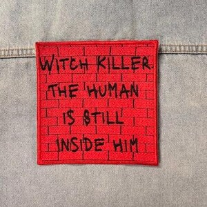May include: A light blue denim jacket featuring a square red patch on the back. The patch has a black brick wall pattern and black embroidered text that reads: WITCH KILLER. THE HUMAN IS STILL INSIDE HIM. The jacket has a collar and seams.