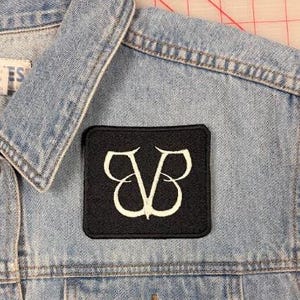 May include: A light blue denim waistcoat featuring a square black embroidered patch with a white stylised logo. The waistcoat has a classic collar, metal buttons, and a chest pocket. A white label with the text L.A. BLUES is visible on the inner collar.