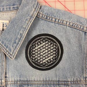 May include: A light blue denim vest featuring a circular black patch with a white embroidered geometric flower of life design. The vest has a classic collar, metal buttons, and a white interior label with text reading L.A. BLUES.