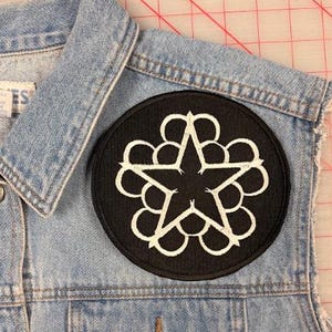 May include: A light blue denim waistcoat featuring a circular black embroidered patch on the chest. The patch displays a white five-pointed star surrounded by a series of interconnected circular loops. The waistcoat has a collar, metal buttons, and a pocket.