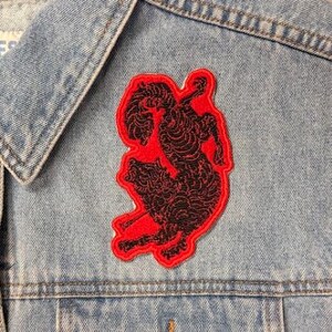 May include: A light blue denim vest featuring a prominent embroidered patch on the chest. The patch displays a black, intricate, swirling line-art design of a rearing horse silhouette, outlined in a vibrant red border. The vest has metal buttons.