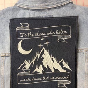 May include: A light blue denim jacket featuring a large rectangular black embroidered patch on the back. The patch displays a white crescent moon, three stars, and mountain peaks with the text "To the stars who listen" and "and the dreams that are answered."