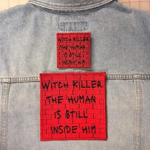 May include: A light blue denim jacket featuring two square red patches with a black brick pattern and the black embroidered text: WITCH KILLER. THE HUMAN IS STILL INSIDE HIM. The patches are positioned vertically on the back of the jacket.