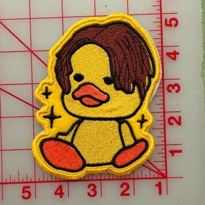 May include: An embroidered patch featuring a yellow duck with brown hair, an orange beak, and orange feet. The duck is surrounded by small black star icons. The patch is positioned on a white cutting mat with red grid lines measuring up to 6 inches.